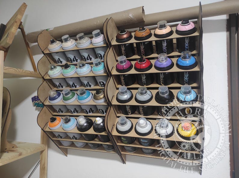 Puede incluir: Estante de almacenamiento de latas de pintura en aerosol de madera lleno de varias latas de pintura en aerosol. Las latas son de diferentes colores, incluyendo rosa, azul y dorado. El estante es de madera marr&oacute;n claro y est&aacute; montado en una pared blanca.