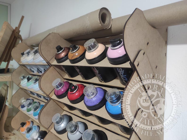 Puede incluir: Un organizador de pintura en aerosol de madera con m&uacute;ltiples compartimentos, que contiene varias latas de pintura en aerosol de colores. Las latas tienen bases negras y tapas de colores. Un rollo de papel est&aacute; montado encima del organizador.