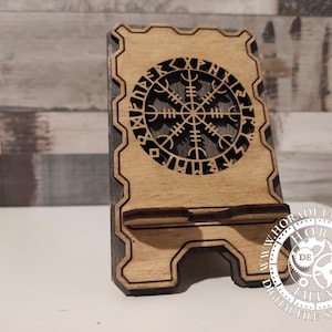 May include: Wooden phone stand with a laser-cut Vegvisir symbol. The stand is light brown with a dark brown border and a slot to hold a phone. The Vegvisir symbol is a Viking compass for guidance.