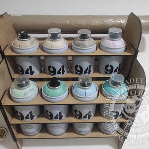 Puede incluir: Un organizador de madera para botes de pintura en aerosol con doce botes. Los botes son blancos con el n&uacute;mero "94" impreso. Las tapas de los botes son de varios colores, incluyendo beige, verde y azul. Un rollo de papel es visible encima del organizador.