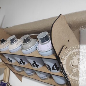 Puede incluir: Un organizador de botes de pintura en aerosol de madera con m&uacute;ltiples compartimentos. El organizador contiene varios botes de pintura en aerosol, cada uno etiquetado con el n&uacute;mero "4". Los botes son blancos con n&uacute;meros negros. El organizador es de madera marr&oacute;n claro.