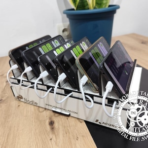 Può includere: Una stazione di ricarica in legno bianco con più slot, ognuno dei quali contiene uno smartphone collegato a un cavo di ricarica bianco. La stazione ha la scritta "Nodle". Una pianta in un vaso blu è sullo sfondo.