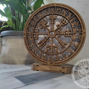 Può includere: Un simbolo Vegvísir in legno, una bussola vichinga, è esposto su un supporto in legno. Il simbolo è intagliato in modo intricato, rivelando uno sfondo più chiaro. Il supporto ha il sito web www.horadetallar.com. Una pianta in vaso è sullo sfondo.