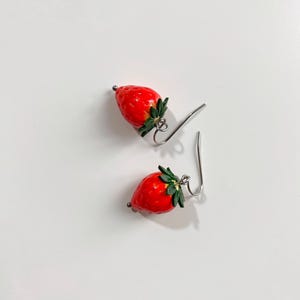 May include: A pair of strawberry earrings. Each earring has a bright red strawberry design with green leaves and a silver-coloured hook. The earrings are set against a white background.