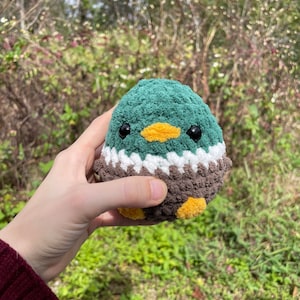 May include: A crocheted plush toy shaped like a duck. The duck is green with a brown chest and yellow beak. It has black eyes and a white stripe around its neck.
