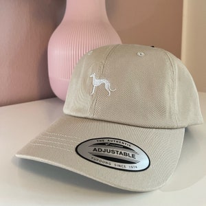 Könnte beinhalten: Eine beige Baseballkappe mit einer weißen gestickten Hundesilhouette auf der Vorderseite. Die Kappe hat einen verstellbaren Riemen mit einem silbernen Etikett, auf dem "The Authentic Yupoong Adjustable Yupoong Since 1974" steht.