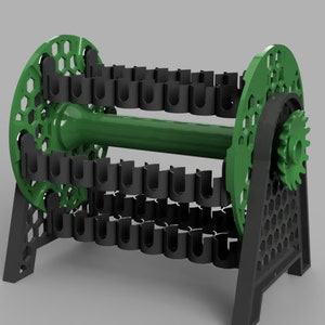 Puede incluir: Un soporte de carrete de plástico verde y negro con un engranaje en un lado. El soporte de carrete tiene un patrón hexagonal en los lados y una serie de ranuras para sujetar carretes.