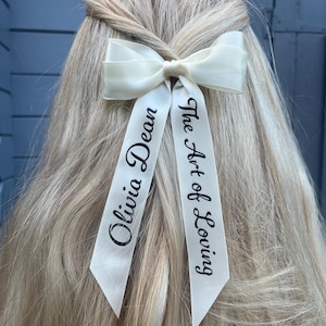 May include: Ivory satin hair bow with long, pointed tails. The tails are printed with the words "Olivia Dean" and "The Art of Loving" in black script. The bow is set against blonde hair.