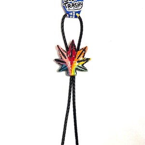 Puede incluir: Un colgante de corbata bolo en forma de hoja de colores del arcoíris con un cordón negro. El colgante está unido a una etiqueta azul que dice "Sew Trashy" en letras blancas y negras. La corbata está sobre un fondo blanco.