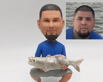 May include: A custom bobblehead figurine of a person wearing a blue shirt and holding a fish. The figurine is made of painted ceramic and has a realistic likeness of the person.