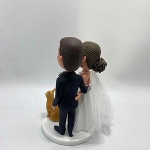 Könnte beinhalten: Ein Hochzeitstortenaufsatz mit einer Braut und einem Bräutigam in traditioneller Hochzeitskleidung, die nebeneinander mit einer kleinen orangefarbenen Katzenfigur stehen. Die Braut trägt ein weißes Kleid mit Schleier, und der Bräutigam trägt einen schwarzen Anzug. Das Paar schaut nach oben, und die Katze schaut das Paar an.