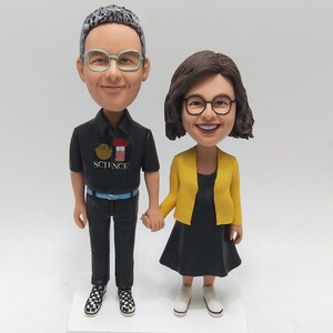 May include: A custom bobblehead figurine set depicting a couple holding hands. The male figure wears a black shirt with the word "SCIENCE" and black trousers. The female figure wears a yellow jacket and black dress. Both figures wear glasses and stand on a white base.