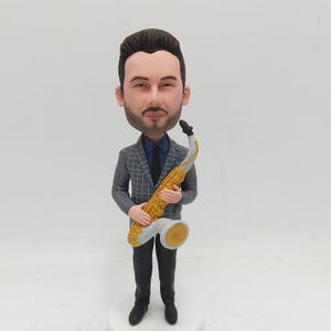 Può includere: Una statuetta personalizzata di una persona che tiene un sassofono dorato. La statuetta indossa una giacca grigia a quadri, una camicia blu e pantaloni neri.