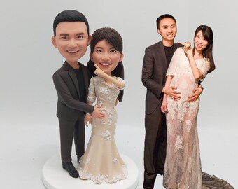 May include: A custom-made figurine couple, the woman in a white lace dress and the man in a black suit. The figurine couple is standing on a white base. In the background, a real-life couple is pictured in a similar pose, the woman in a white lace dress and the man in a black suit.