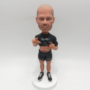 Könnte beinhalten: Eine Bobblehead-Figur eines glatzköpfigen Mannes, der ein schwarzes T-Shirt mit dem Namen "ELLIOTT" und schwarze Shorts mit dem Wort "PSYCHO" trägt. Die Figur steht auf einer weißen, kreisförmigen Basis und trägt schwarze Turnschuhe.
