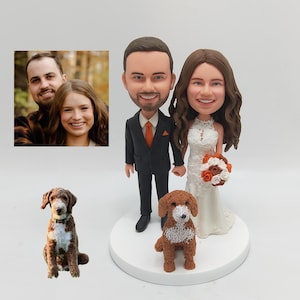 May include: A wedding cake topper featuring a bride and groom in a cartoon style, holding hands. The bride is wearing a white dress and the groom is wearing a black suit with an orange tie. A brown and white dog is standing in front of them.