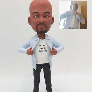 Op de afbeelding: Een gepersonaliseerd beeldje van een persoon met een kale kop en een baard, die een lichtblauw shirt en een zwarte broek draagt. Het shirt is open om een wit T-shirt met de tekst "I Love North Carolina" te laten zien.