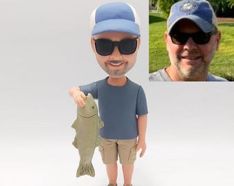 May include: A custom bobblehead figurine of a person wearing a blue and white baseball cap, sunglasses, a blue t-shirt, khaki shorts, and green shoes. The person is holding a fish.