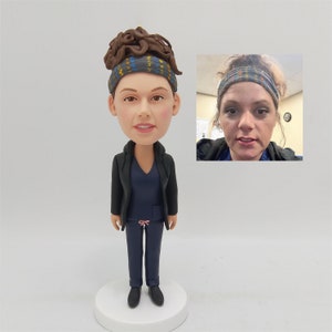 Op de afbeelding: Een gepersonaliseerd bobblehead-beeldje van een persoon die een zwart jack, blauwe scrubs en een geprint hoofdband draagt. Het beeldje staat op een witte basis. Het gezicht van de persoon is zichtbaar en heeft bruin haar.