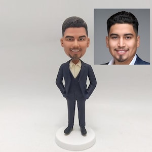May include: A custom bobblehead figurine of a person wearing a dark blue suit with a white shirt and a light yellow vest. The figurine is standing on a white base.