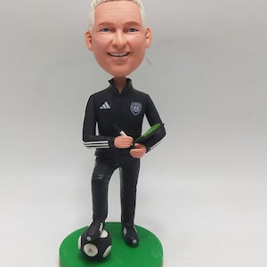 May include: A bobblehead figurine of a soccer coach in a black Adidas tracksuit, holding a pen and notepad. The figurine stands on a green base with a soccer ball. The coach has white hair and is smiling.