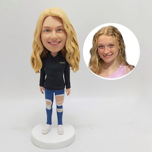 Op de afbeelding: Een op maat gemaakte bobblehead-figuur van een persoon met blond krullend haar, gekleed in een zwarte hoodie en gescheurde blauwe jeans. Het beeldje staat op een witte basis, met een kleinere afbeelding van de persoon in een cirkel.