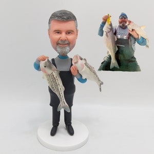 Może przedstawiać: Spersonalizowana figurka osoby trzymającej dwie ryby. Figurka jest wykonana z malowanej ceramiki i ma na sobie niebieską koszulę, czarne spodnie i czarny fartuch. Osoba ma siwą brodę i siwe włosy. Figurka stoi na białej podstawie.