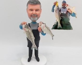 May include: A custom figurine of a person holding two fish. The figurine is made of painted ceramic and is wearing a blue shirt, black pants, and a black apron. The person has a gray beard and gray hair. The figurine is standing on a white base.