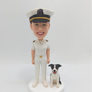 Op de afbeelding: Een gepersonaliseerd bobblehead-beeldje van een persoon in een wit marineuniform en een zwarte hoed met gouden versieringen. Het beeldje staat naast een kleine zwart-witte hond.