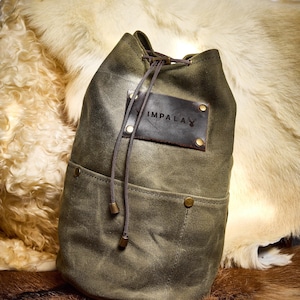 Peut inclure: Un sac à cordon en toile verte avec un patch en cuir sur lequel est inscrit "IMPALA" et un logo de tête de cerf. Le sac a une grande poche sur le devant et est posé sur une surface en fourrure brune.