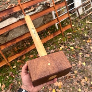 May include: A wooden mallet with a light-coloured handle and a dark brown head. The handle has the word "IMPALAY" printed on it. The head has a circular brass accent. The mallet is held in a hand.