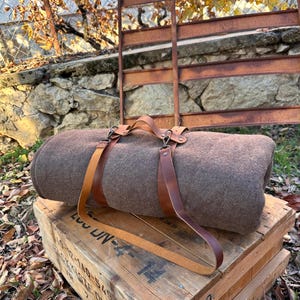 Puede incluir: Una manta de lana marrón enrollada con correas y un asa de cuero marrón. La manta descansa sobre una caja de madera con marcas descoloridas. El fondo presenta una verja de metal oxidado y un muro de piedra.