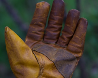 May include: A brown leather work glove with a reinforced palm and fingers. The glove has a cuff with a snap closure.