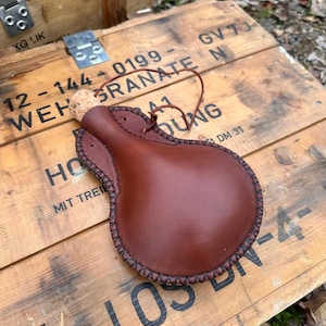 May include: A brown leather flask with a cork stopper and a stitched border. The flask has a leather strap and is resting on a wooden crate. The crate has text printed on it.