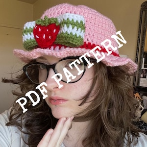 Op de afbeelding: Een roze gehaakte hoed met een groene en witte gehaakte aardbeien strik. De hoed heeft een brede rand en is gemaakt van zacht garen. De tekst "PDF PATTERN" is over de afbeelding gelegd.
