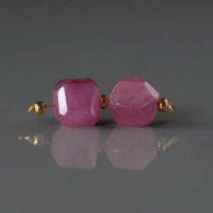 May include: A pair of faceted, pink gemstone earrings. The gemstones are a vibrant magenta color and are connected by small gold-colored posts. The earrings are displayed on a reflective surface.