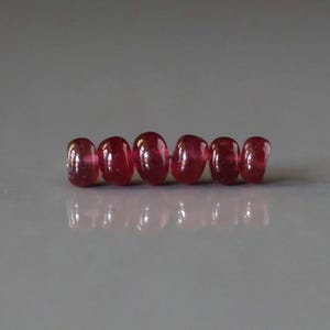 May include: Six polished, oval-shaped ruby gemstones. The rubies are a deep, translucent red color, with a smooth, glossy surface. They are arranged in a row, reflecting light on a gray surface. Ideal for jewelry making or collecting.