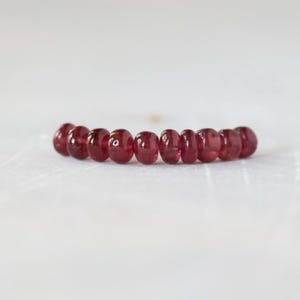 May include: A ring featuring a single row of ten small, round, translucent deep red beads. The beads are arranged in a horizontal line, each displaying a smooth, polished surface with subtle variations in color depth and light reflection.