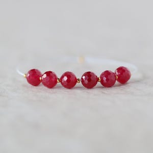 May include: A bracelet featuring six faceted, round, deep red beads arranged in a row on a thin, white cord. Small, metallic gold-toned spacer beads are positioned between each of the red beads, creating a structured and symmetrical design.