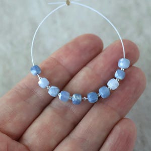 May include: A strand of nine faceted, light blue cube-shaped beads separated by small silver-toned spacer beads. The beads are strung on a thin, flexible white wire that forms a circular loop, showcasing a gradient of soft blue tones.