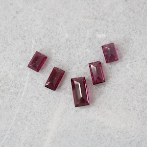 May include: Five deep magenta, rectangular baguette-cut gemstones arranged in a slight arc. Each stone features faceted surfaces that reflect light, showcasing a translucent, saturated purple-red color and a clean, sharp geometric shape.