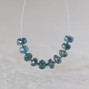 May include: A strand of nine faceted, round, blue-green gemstone beads alternating with small, smooth, gold-toned metal spacer beads. The beads are strung on a thin, clear filament, arranged in a curved, U-shaped formation against a neutral background.