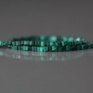 May include: Close-up of a necklace made of small, square, malachite beads. The beads are a deep green color, with variations in shade. The necklace is displayed on a reflective surface, creating a mirrored effect. The beads are strung together, forming a delicate piece of jewelry.