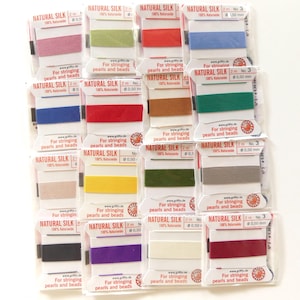 May include: A collection of spools of Natural Silk thread in various colors, including pink, green, blue, red, and yellow. Each spool is labeled with text indicating its use for stringing pearls and beads. The packaging is clear plastic.