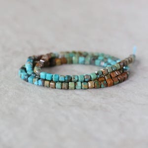 May include: A strand of small, faceted cube-shaped beads in varying shades of turquoise blue and earthy brown. The beads are arranged in a circular coil, showcasing a mix of bright blue tones and darker, mottled brown textures throughout the strand.