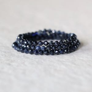 May include: A multi-strand bracelet composed of small, faceted, deep navy blue beads. The beads are arranged in three distinct circular rows, creating a layered appearance with a reflective, textured surface throughout the entire piece.