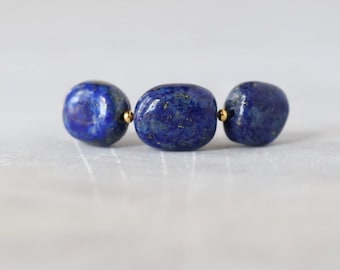 Könnte beinhalten: Drei polierte, oval geformte Lapislazuli-Perlen in tiefem Blau mit natürlichen goldfarbenen Sprenkeln. Die Perlen sind in einer horizontalen Reihe angeordnet und durch kleine, runde, metallisch goldfarbene Zwischenperlen getrennt.