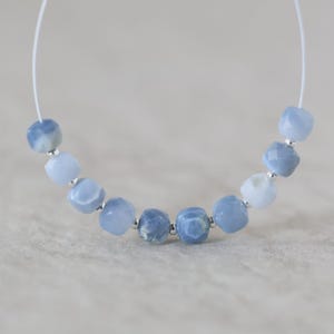 May include: A strand of nine faceted, light blue, semi-translucent beads arranged in a curved line. Each bead is separated by a small, metallic silver spacer bead, and the entire set is strung on a thin, white, flexible cord for jewelry making.