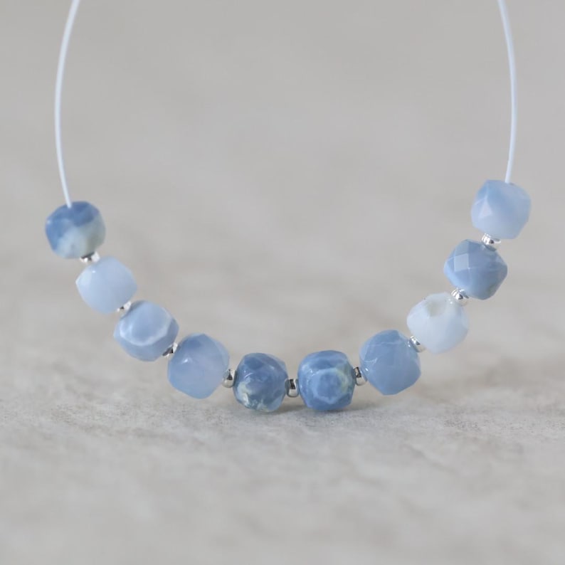 May include: A strand of nine faceted, light blue, semi-translucent beads arranged in a curved line. Each bead is separated by a small, metallic silver spacer bead, and the entire set is strung on a thin, white, flexible cord for jewelry making.