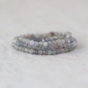 May include: A multi-strand bracelet composed of small, faceted, translucent beads in muted shades of gray, blue, and soft lavender. The beads are strung together in a circular arrangement, showcasing a reflective, textured surface on each individual stone.